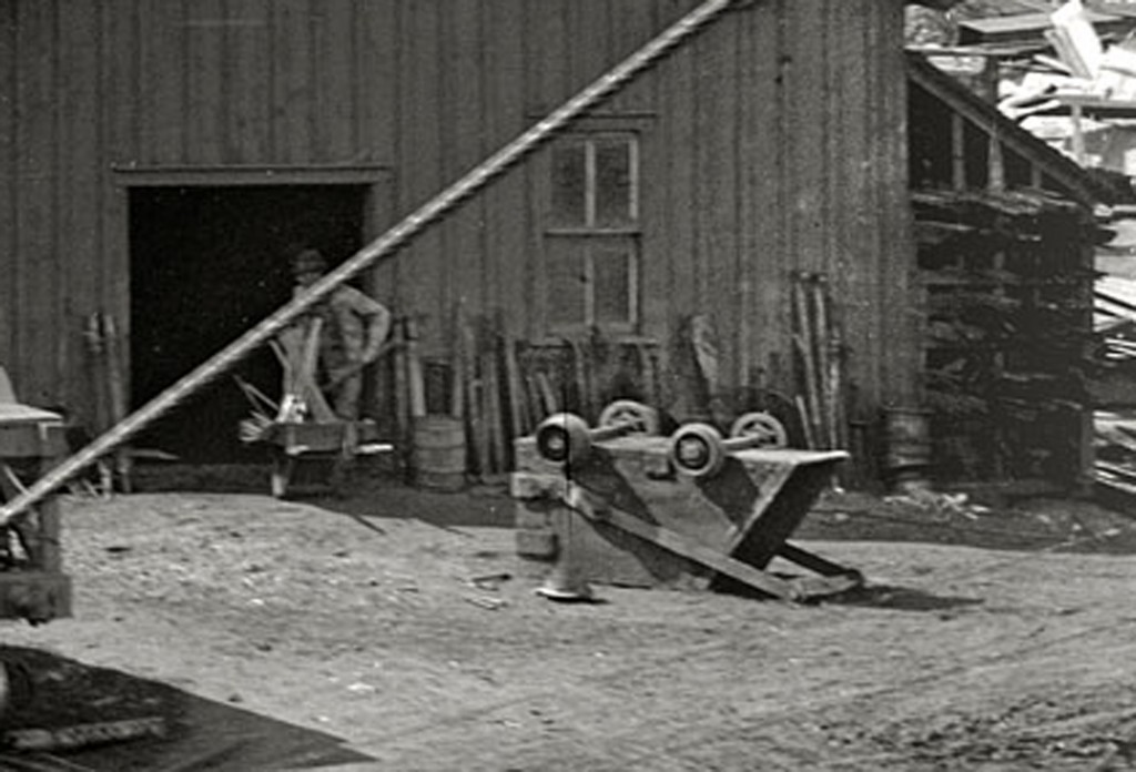 This building appears to be a repair or blacksmith shop. Materials and tools are stacked against the building. A skip is upside down, presumably to be fixed. It appears the ore cars may need repairs to. The car closest is only partially filled while the other two are not.